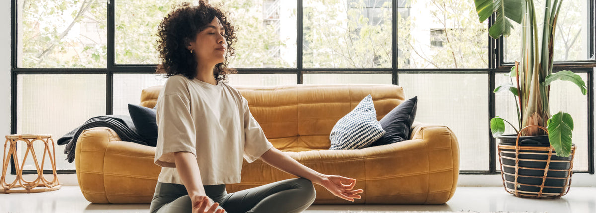 A woman with her eyes closed sits cross-legged on a rug and meditates in a sunny living room